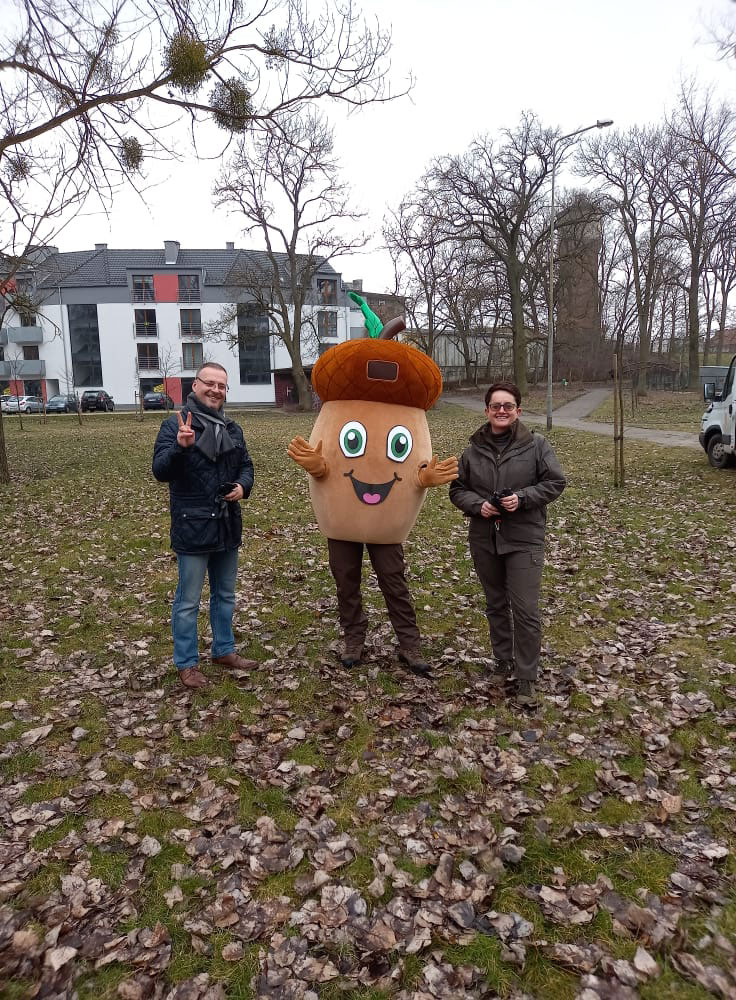 Na fotografii widnieje kostium w postaci żołędzia, gdzieś w plenerze, w towarzystwie dwóch osób. W tle widoczne drzewa i budynek